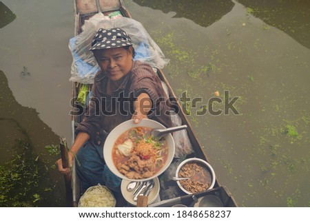 Market woman giving noodle at Damnoen Saduak Floating Market or Amphawa. Local people sell fruits, traditional food on boats in canal, Ratchaburi District, Thailand. Famous Asian tourist attraction.