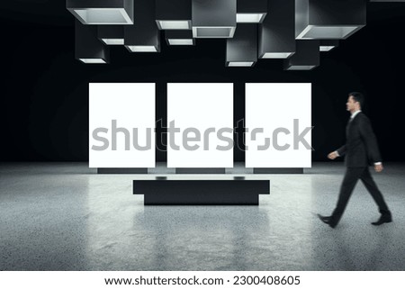 Marketing concept with businessman walking by three blank white digital screens with place for advertising or logo brand in abstract industrial dark hall with concrete floor and background, mock up