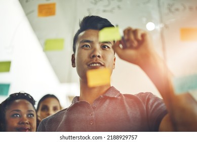 A marketing professional brainstorming ideas with colleagues, writing on transparent board with sticky notes during meeting. Young, Asian entrepreneur discussing a work project schedule or timeline. - Shutterstock ID 2188929757