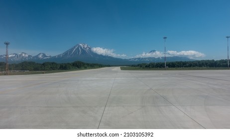 Marking lines and tire tracks are visible on the concrete runway of the airfield. Snow-capped conical volcanoes against a background of blue sky and clouds. Copy space. Petropavlovsk-Kamchatsky