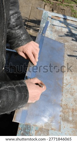 Marking out and measuring the required dimensions on a small sheet of metal lying on a work surface in an outdoor craft workshop, using a pencil and ruler in the hands of a craftsman marking out the iron plate.