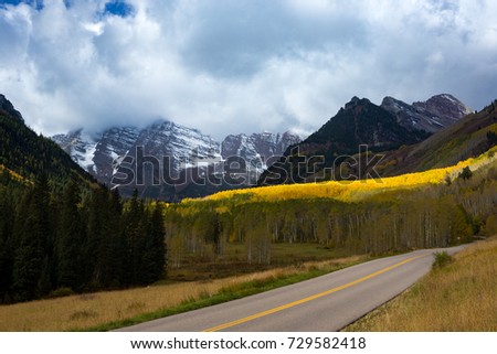 Maroon Creek Road in a glacial valley along with pines and yellow gold aspen trees in fall foliage autumn season with cloudy Maroon Peak and Maroon North Peak in background, Pitkin County,Colorado USA