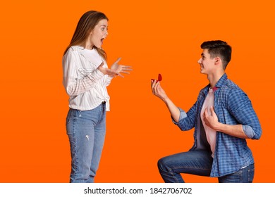 Marriage proposal and big surprise. Smiling young man kneeling and giving ring in box, shocked woman screams with joy and look at male, isolated on orange background, studio shot, empty space