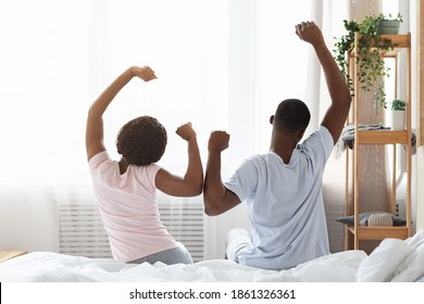 Married black couple waking up in morning, sitting on bed, stretching in cozy bedroom, back view, free space. Unrecognizable african american family starting day together, raising hands up