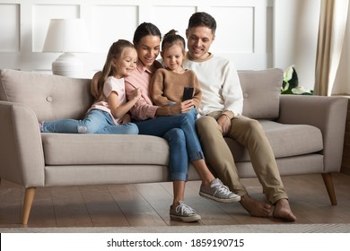 Married couple with 2 preschool adorable daughters resting seated on couch in living room with modern gadget having fun using smartphone. Take family picture, make videocall, parental control concept - Powered by Shutterstock