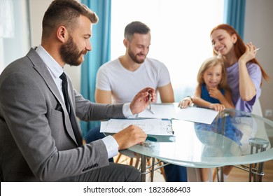 Married Couple With Child Girl Going To Sign The Documents For Getting Their First Apartment, They Sit With Real Estate Agent, Male In Suits Ready To Give Keys To Family