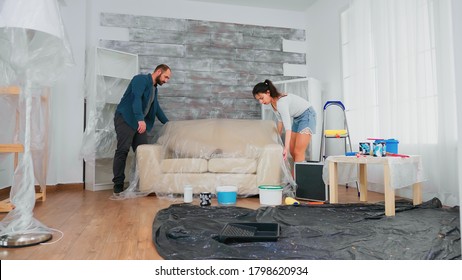 Married Couple Covering Sofa With Plastic Sheet For Home Decorating. Apartment Redecoration And Home Construction While Renovating And Improving. Repair And Decorating.