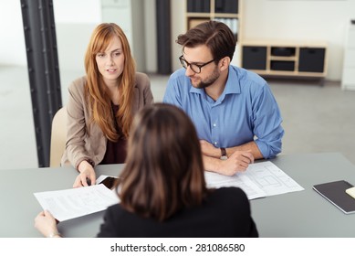 Married couple discussing investments with a broker as they sit together at a desk in her office going through paperwork together, view from behind the agent