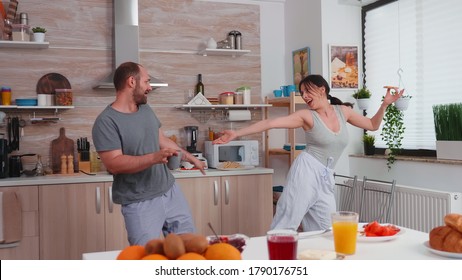 Married Couple Having Fun Dancing In Kitchen During Breakfast. Carefree Husband And Wife Laughing, Singing, Dancing Listening Musing, Living Happy And Worry Free. Positive People.