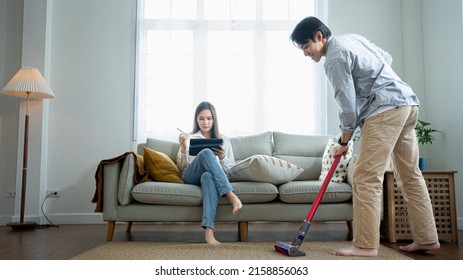 Married Couple Holiday Asian American Man Doing House Work Lazy Wife Resting On Sofa Chatting Online Enjoy Free Time Weekend While Thai Husband Doing Chores Using Vacuum Cleaner Cleaning Carpet Home.