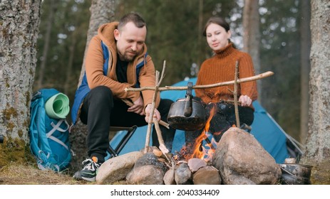 A Married Couple Prepares Food On A Campfire On The Shore Of The Lake With A Tent. Hiking Camp With A Bonfire In The Forest. A Man And A Woman Enjoy The Silence By The Fire Near The Tent