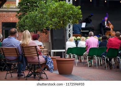 Married Couple Is Sitting In Restaurant, Drinking Wine, Eating Fancy Snacks And Listening To Jazz Concert In Concert Venue In City Square. Musicians, Stage, Crowd And Concert In The Background.
