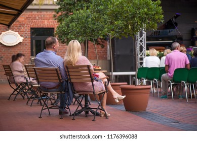 Married Couple Is Sitting In Restaurant, Drinking Wine, Eating Fancy Snacks And Listening To Jazz Concert In Concert Venue In City Square. Musicians, Stage, Crowd And Concert In The Background.
