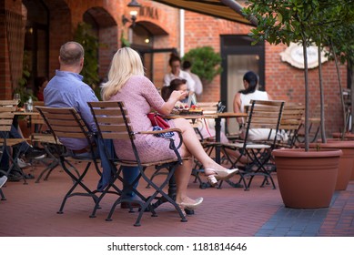 Married Couple Is Sitting In Restaurant, Drinking Wine, Eating Fancy Snacks And Listening To Jazz Concert In Concert Venue In City Square. Musicians, Stage, Crowd And Concert In The Background.
