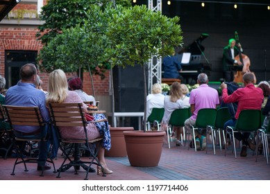 Married Couple Is Sitting In Restaurant, Drinking Wine, Eating Fancy Snacks And Listening To Jazz Concert In Concert Venue In City Square. Musicians, Stage, Crowd And Concert In The Background.
