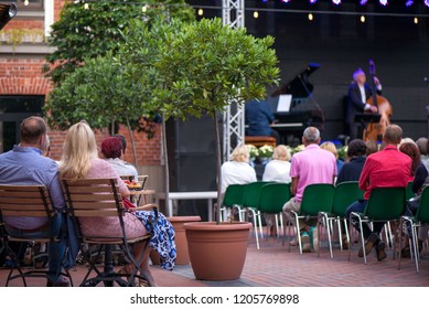 Married Couple Is Sitting In Restaurant, Drinking Wine, Eating Fancy Snacks And Listening To Jazz Concert In Concert Venue In City Square. Musicians, Stage, Crowd And Concert In The Background.
