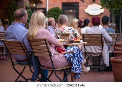 Married Couple Is Sitting In Restaurant, Drinking Wine, Eating Fancy Snacks And Listening To Jazz Concert In Concert Venue In City Square. Musicians, Stage, Crowd And Concert In The Background.
