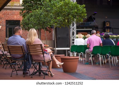 Married Couple Is Sitting In Restaurant, Drinking Wine, Eating Fancy Snacks And Listening To Jazz Concert In Concert Venue In City Square. Musicians, Stage, Crowd And Concert In The Background.
