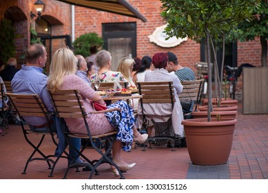 Married Couple Is Sitting In Restaurant, Drinking Wine, Eating Fancy Snacks And Listening To Jazz Concert In Concert Venue In City Square. Musicians, Stage, Crowd And Concert In The Background.
