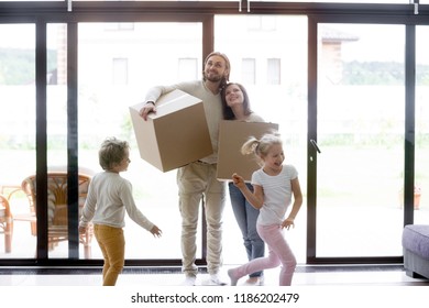 Married Couple Standing In Living Room With Little Children. Family Moving At New House. Smiling Parents Holding Unopened Cardboard Boxes, Joyful Preschool Son And Daughter Running And Playing Around