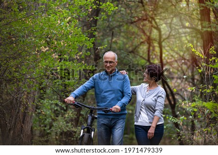 Married couple in their sixties take a leisure walk in the green forest with a bike; young feeling Caucasian husband and wife smiling and talking