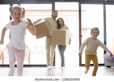 Married Millennial Couple Enter Living Room With Little Children. Family Moving At New House. Smiling Parents Holding Cardboard Boxes, Joyful Toddler Son And Preschool Daughter Running Playing Around