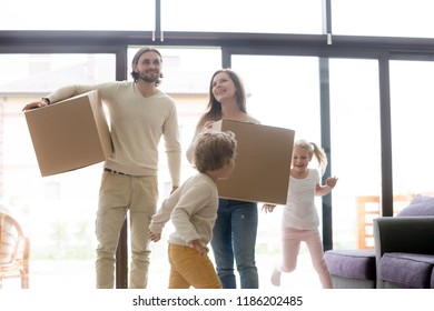 Married Millennial Couple Enter Living Room With Little Children. Family Moving At New House. Young Smiling Parents Holding Cardboard Boxes, Joyful Toddler Son And Daughter Running And Playing Around