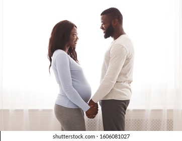 Married pregnant afro couple holding hands and looking at each other, staying next by window