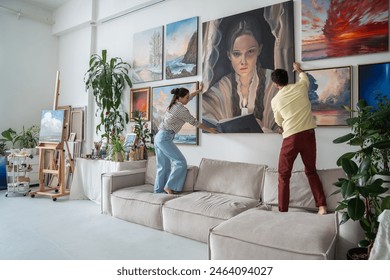 Married talented couple hangs trendy art canvas on wall in spacious bright living room gallery. Focused woman artist with man architect in home art studio discussion new art exhibition. Art lovers.  - Powered by Shutterstock