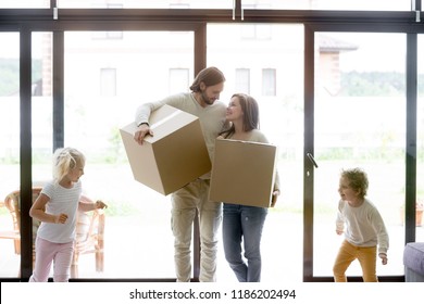 Married Young Couple Enter Living Room With Little Children. Family Moving At New House. Smiling Parents Holding Unopened Cardboard Boxes, Joyful Toddler Son And Daughter Running And Playing Around