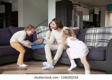 Married Young Couple With Little Children Spend Free Time In Living Room At Wealthy New Home. Smiling Parents Sits On Couch And Playing With Preschool Son And Daughter. Happy Family At House Concept