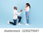 Marry me. Black middle aged man holding giving open box with engagement ring to excited young woman, asking her to be his wife during romantic date standing on one knee, blue studio background