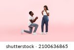 Marry Me. Happy black man holding giving open box with engagement ring to excited girlfriend asking her to be his wife during romantic date standing on one knee, pink studio wall, banner, copy space