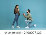 Marry Me. Happy indan man holding giving open box with engagement ring to excited girlfriend asking her to be his wife during romantic date standing on one knee, blue studio wall, banner, copy space