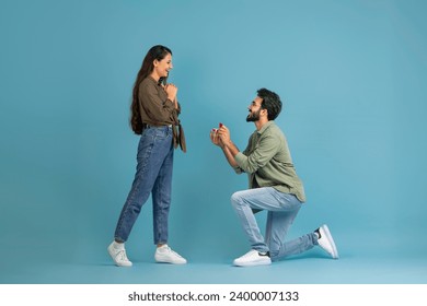 Marry Me. Happy indan man holding giving open box with engagement ring to excited girlfriend asking her to be his wife during romantic date standing on one knee, blue studio wall, banner, copy space