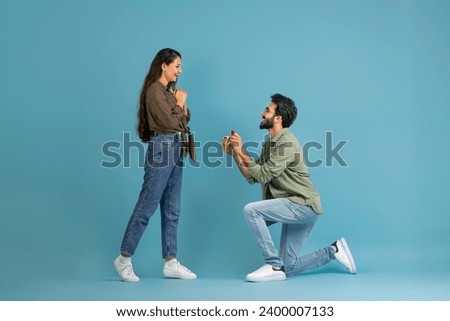 Marry Me. Happy indan man holding giving open box with engagement ring to excited girlfriend asking her to be his wife during romantic date standing on one knee, blue studio wall, banner, copy space Foto d'archivio © 