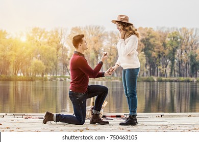 Marry me. Horizontal shot of young man in casual clothing standing on one knee and holding engagement ring while making wedding proposal to his beautiful girlfriend. Engagement of young couple.