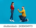 Marry Me. Romantic young man making proposal to his girlfriend, loving millennial guy kneeling and presenting engagement ring to surprised woman, capturing heartfelt moment against blue background