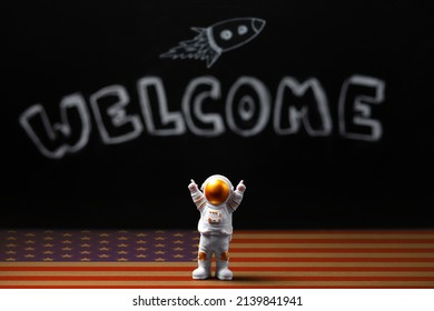 Mars colonization. Figure of astronaut with victoriously raised hands. In the background, a chalky black board with the inscription welcome and a flying rocket, a floor with an American flag design.