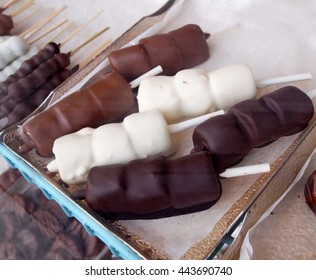 Marshmallow Candy Enrobed In Dark Chocolate, White Chocolate, And Milk Chocolate, On Sticks On A Tray,  In A Glass Display Case At A Candy Shop.