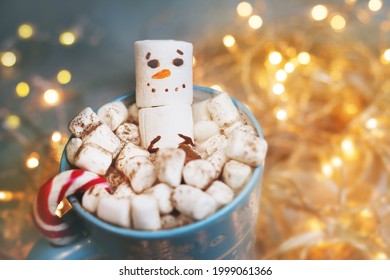 Marshmellow Smiling Snowman In Winter Mug Against Background Of Blur Festive Yellow Lights And Glow