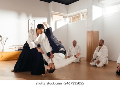 A martial arts instructor skillfully performs an aikido throw on a student during a demonstration, with attentive practitioners observing from the mat. Text: 