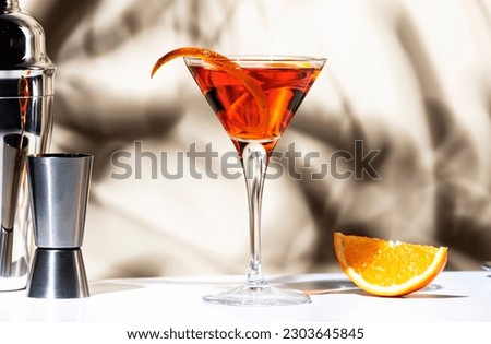 Martinez orange cocktail drink with red vermouth, liqueur, bitter, citrus zest and ice in martini glass. Light beige background, hard light, shadow pattern