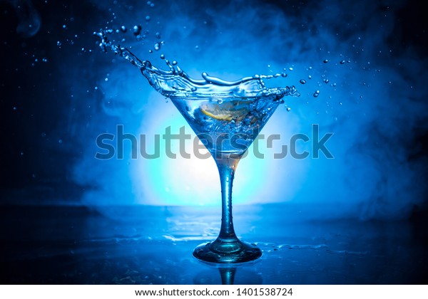 Martini\
cocktail glass splashing on dark toned smoky background or colorful\
cocktail in glass with splashes and lemon. Party club\
entertainment. Mixed light. Selective\
focus