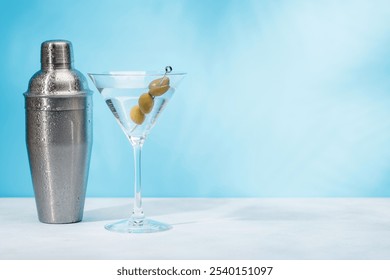 A martini cocktail and cocktail shaker arranged on a table against a light blue background, enhanced by sunny glares and playful shadows - Powered by Shutterstock