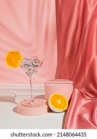 Martini glass with ice and orange fruit, palm leaf shadow and podium on pink background. Pastel pink silk curtain. Summer drink minimal concept. Suitable for Product Display and Business Concept.