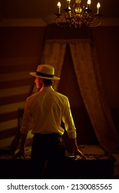 A Masculine Brunet Man In A Classic White Shirt And A Hat Stands With His Back Turned In A Dark Bedroom Near A Table With Playing Cards And Banknotes. Detective Story, Mafia, Gambling.