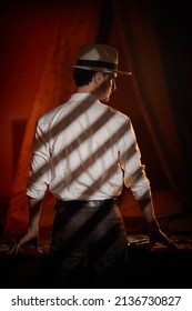 A Masculine Brunet Man In A Classic White Shirt And Hat Stands With His Back To The Camera In A Dark Bedroom Near A Table With Playing Cards And Banknotes. Detective Story, Mafia, Gambling.