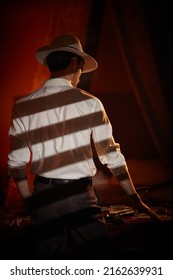 A Masculine Brunet Man In A Classic White Shirt And A Hat Stands With His Back Turned In A Dark Bedroom Near A Table With Playing Cards And Banknotes. Detective Story, Mafia, Gambling.