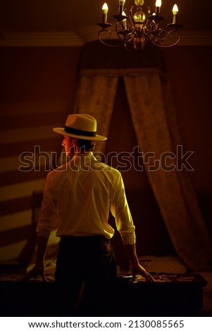 A masculine brunet man in a classic white shirt and a hat stands with his back turned in a dark bedroom near a table with playing cards and banknotes. Detective story, mafia, gambling.